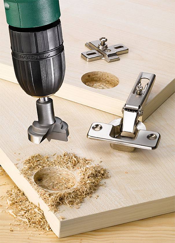 Bohrmaschine mit Forstnerbohrer erzeugt ein präzises Loch in Holz; daneben liegt ein Scharnier zur Montage.