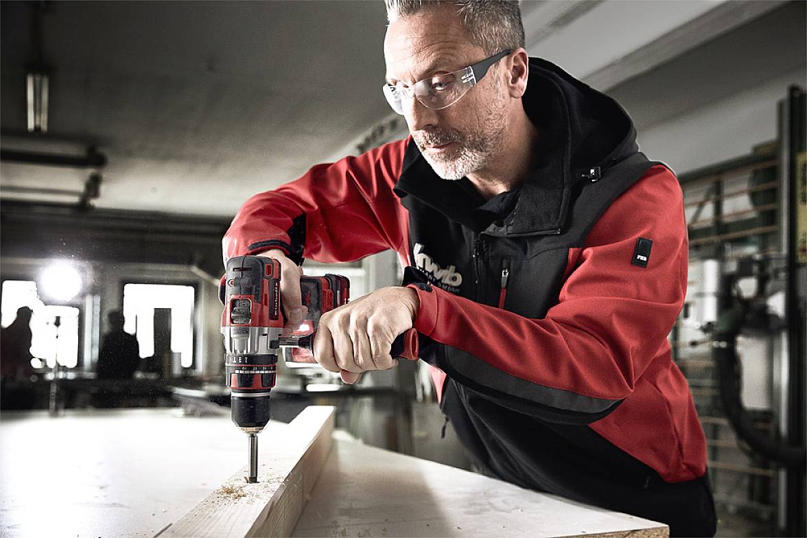 Ein Mann in Schutzbrille und roter Arbeitsjacke bohrt mit einer Bohrmaschine ein Loch in ein Holzbrett in einer Werkstatt.