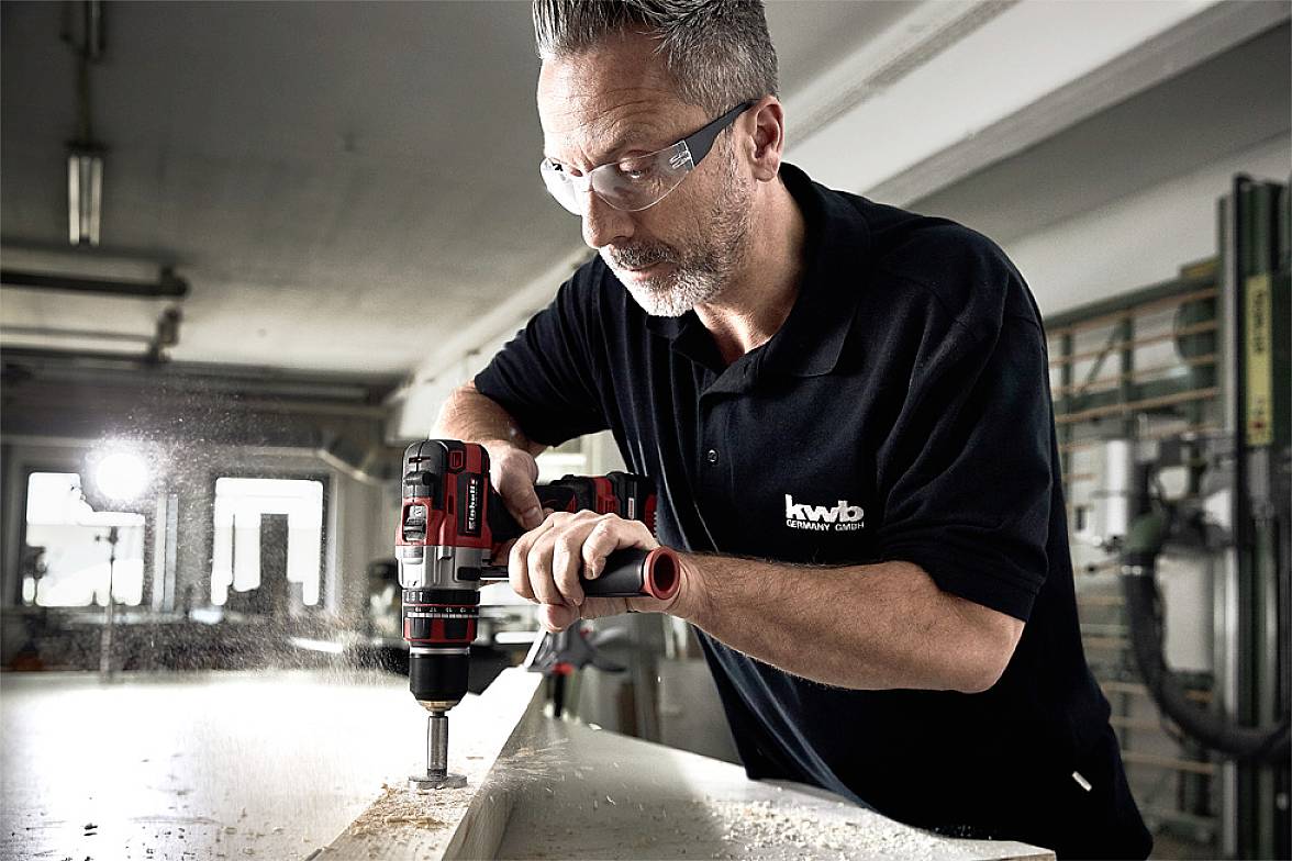 Ein Mann trägt Schutzbrille und benutzt eine Bohrmaschine, um Holz zu bearbeiten. Er arbeitet konzentriert in einer Werkstattumgebung.