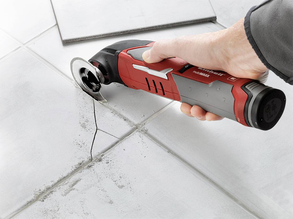 Eine Hand hält ein oszillierendes Werkzeug mit rotem Griff, schneidet eine Fliese entlang eines Risses auf einem weißen Fliesenboden.