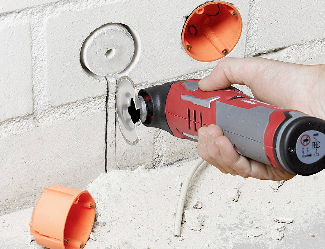 Person verwendet ein rot-graues Werkzeug, um Löcher in eine weiße Ziegelwand zu schneiden, mit sichtbaren orangefarbenen Elektroinstallationsdosen.