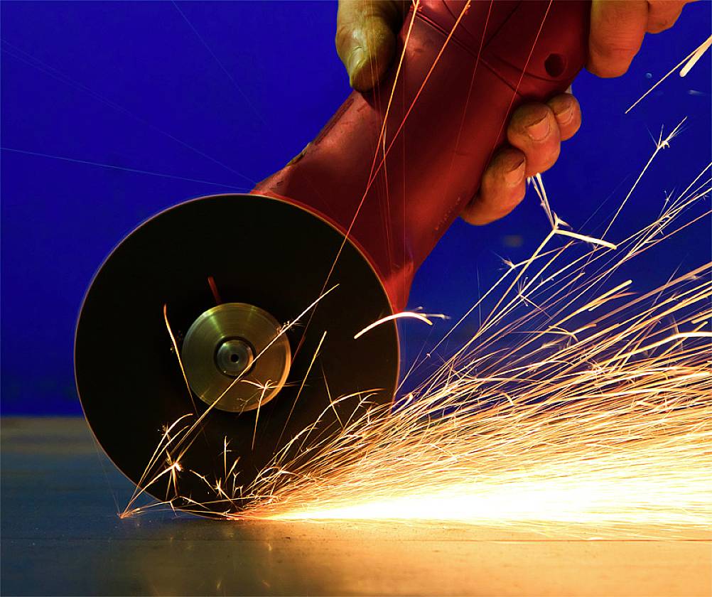 Eine Hand hält einen Winkelschleifer, der Funken sprüht, während er Metall bearbeitet. Hintergrund ist blau.