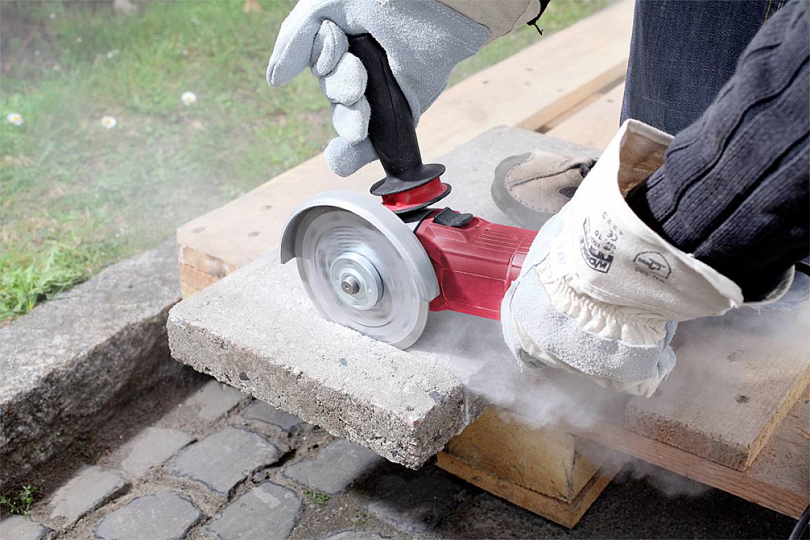 Eine Person schneidet mit einer roten Winkelschleifmaschine Beton. Staub steigt auf, während der Schnitt auf einem Holzbalken erfolgt.