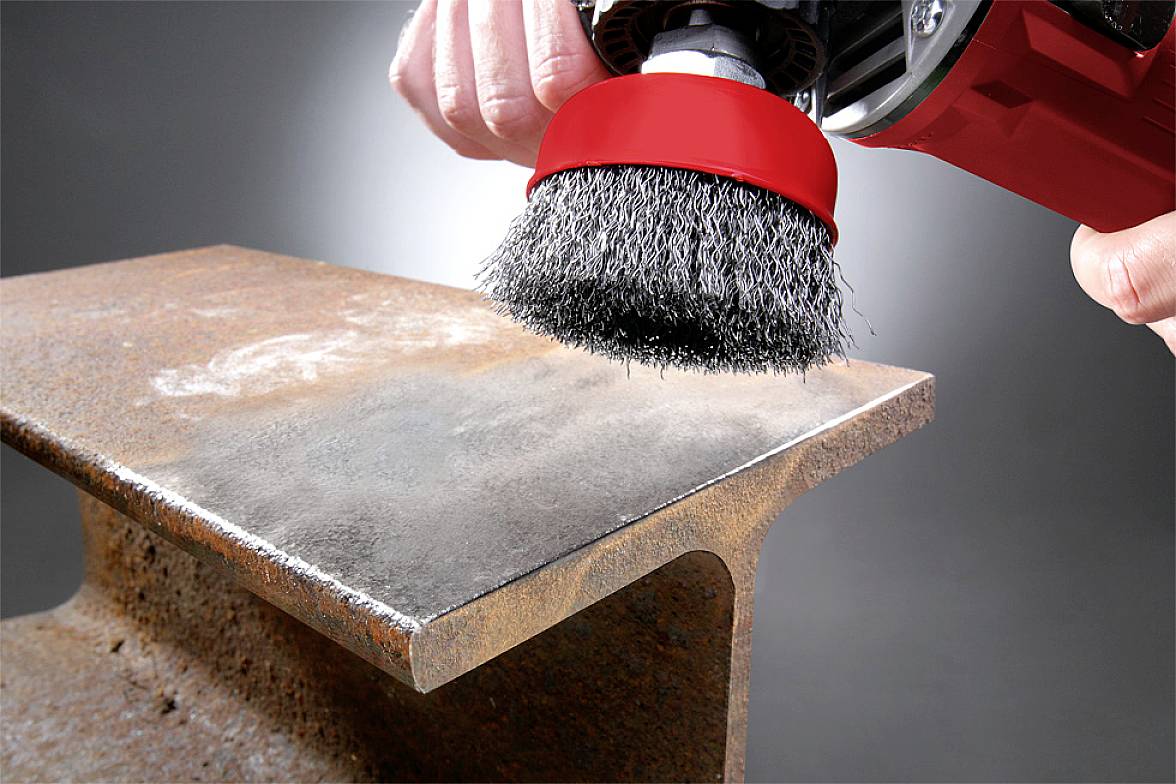 Eine Drahtbürste entfernt Rost von einem Metallträger. Hände halten das Gerät sicher. Der Träger zeigt deutliche Korrosionsspuren.