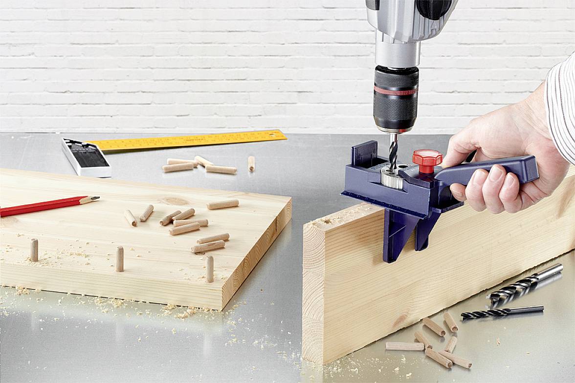 Eine Hand hält eine Bohrmaschine und bohrt Löcher in ein Holzbrett. Auf dem Tisch liegen Dübel, ein Maßstab, ein Bleistift und Bohrer.