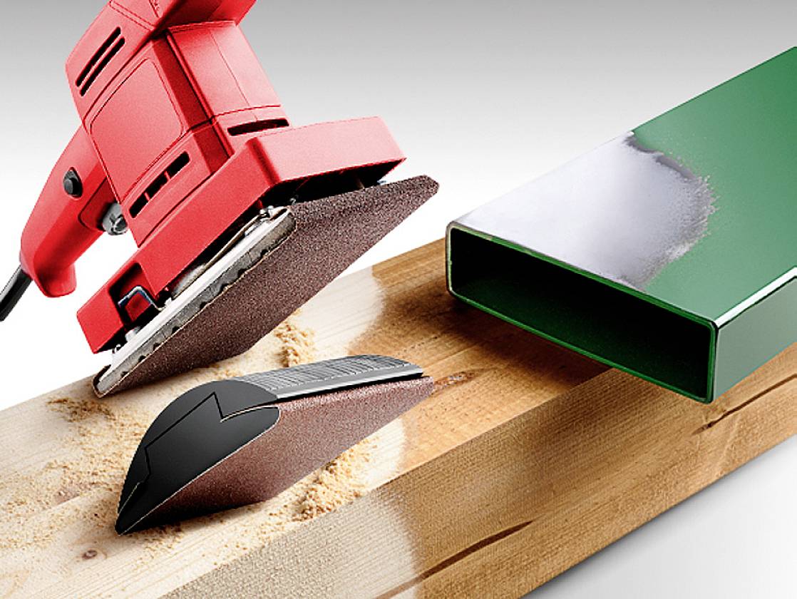 Ein rotes Schleifgerät schleift Holz, ein kleines schwarzes Schleifwerkzeug liegt daneben. Rechts ist ein teilweise lackiertes Metallrohr.