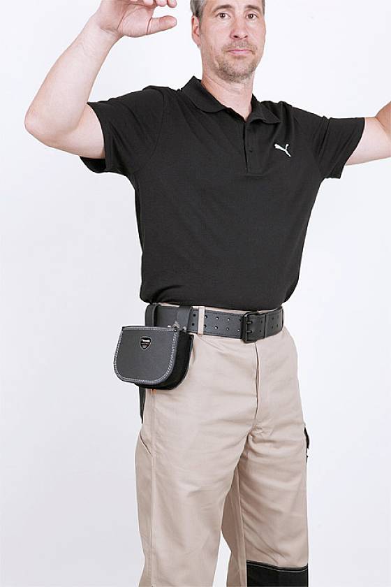 Ein Mann in einem schwarzen Polohemd und beigen Hosen hebt die Arme. Er trägt eine schwarze Gürteltasche.