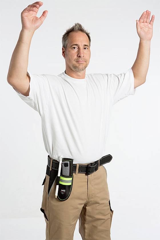 Ein Mann in beiger Hose und weißem T-Shirt hebt beide Arme. An seinem Gürtel ist ein Gerät befestigt. Hintergrund ist weiß.