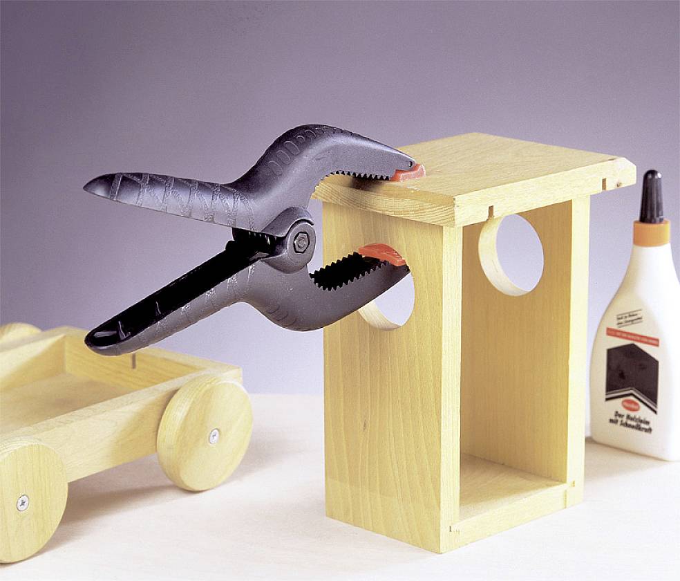 Holzbausatz mit einem F-Klemmwerkzeug, das zwei Teile zusammenhält. Daneben steht eine Flasche Holzleim; links liegt ein Holzspielzeugauto.