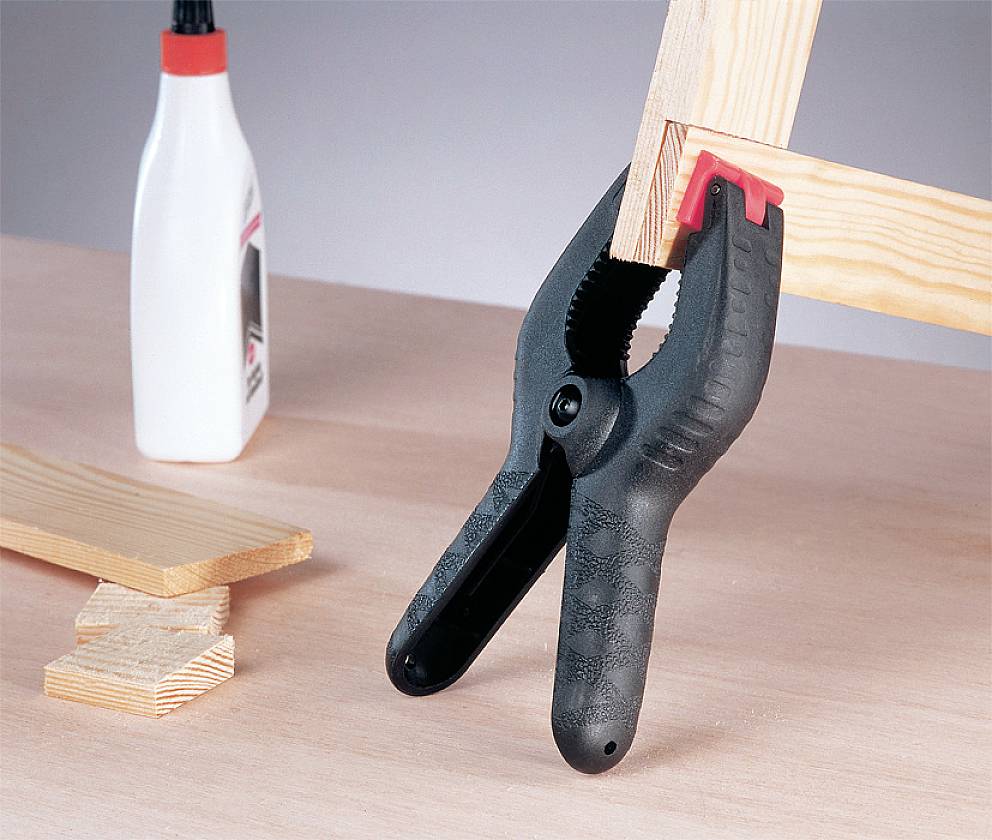 Eine schwarze Federzwinge hält zwei Holzstücke zusammen, daneben stehen eine Flasche Holzleim und kleine Holzreste auf einem Tisch.