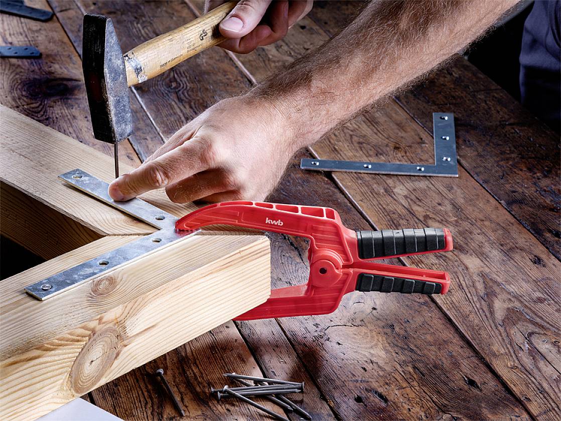 Eine Person hämmert einen Nagel in ein Holzstück, das mit einer roten Klemme fixiert ist. Ein Winkelmesser liegt daneben auf dem Tisch.