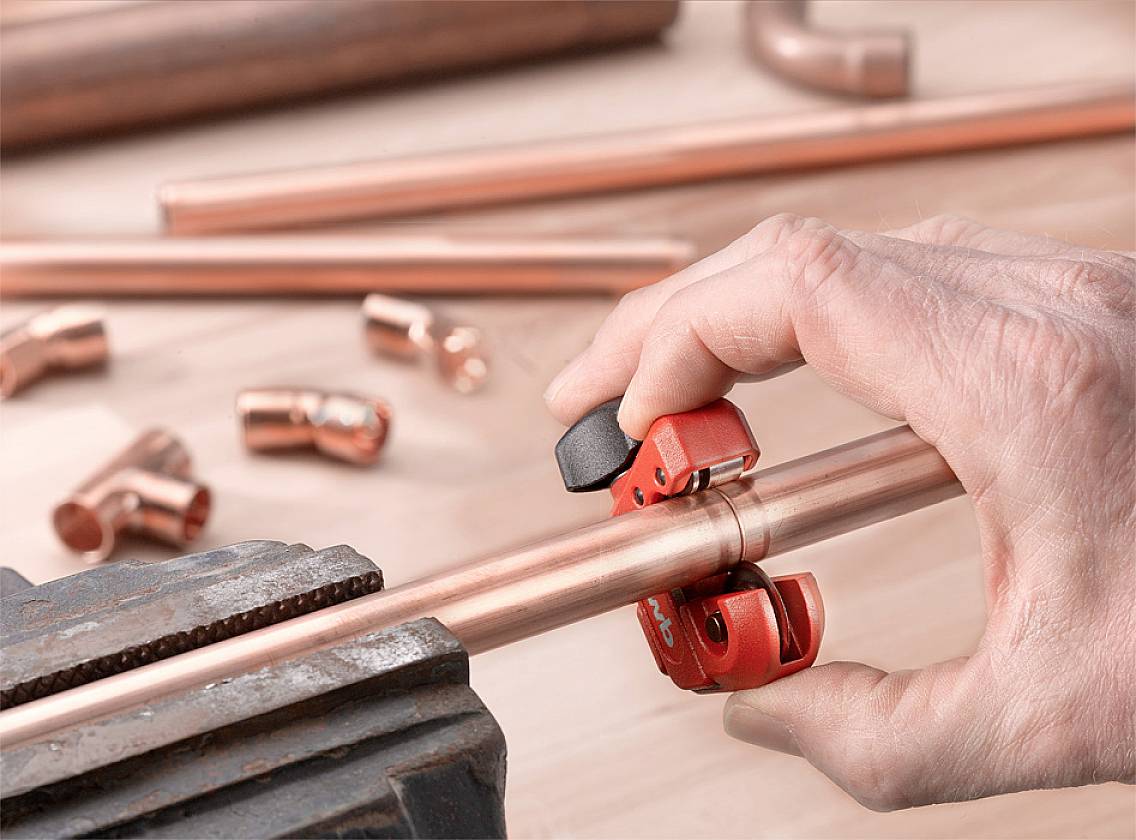 Eine Hand schneidet ein kupfernes Rohr mit einem Rohrschneider, umgeben von weiteren Rohrstücken und Fittings auf einem Holztisch.