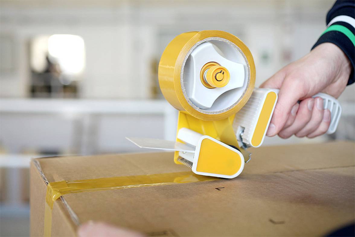 Eine Person verwendet einen gelben Handabroller, um ein braunes Paket mit transparentem Klebeband zu versiegeln.