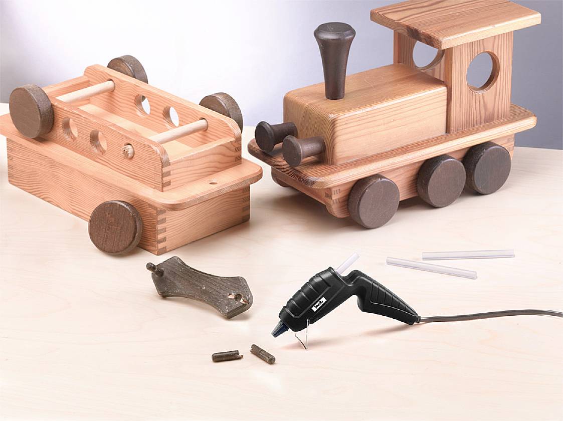 Holzspielzeug in Form eines Zuges und Wagens auf einem Tisch. Daneben liegt eine Heißklebepistole mit Klebestiften und kleinen Holzstücken.