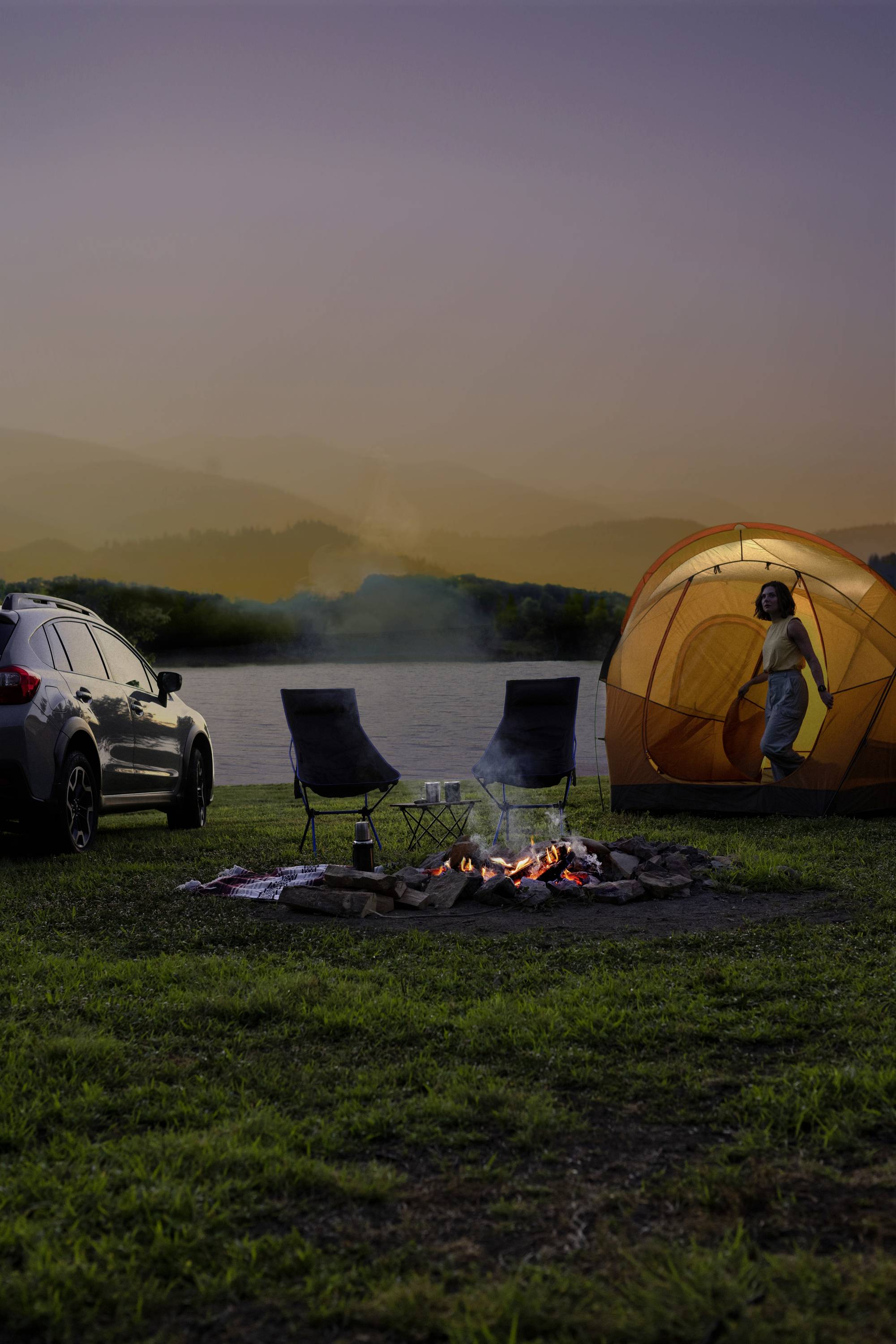 Auto neben Zelt am Seeufer bei Sonnenuntergang. Zwei Stühle und Lagerfeuer im Vordergrund, Person schaut aus dem Zelt.