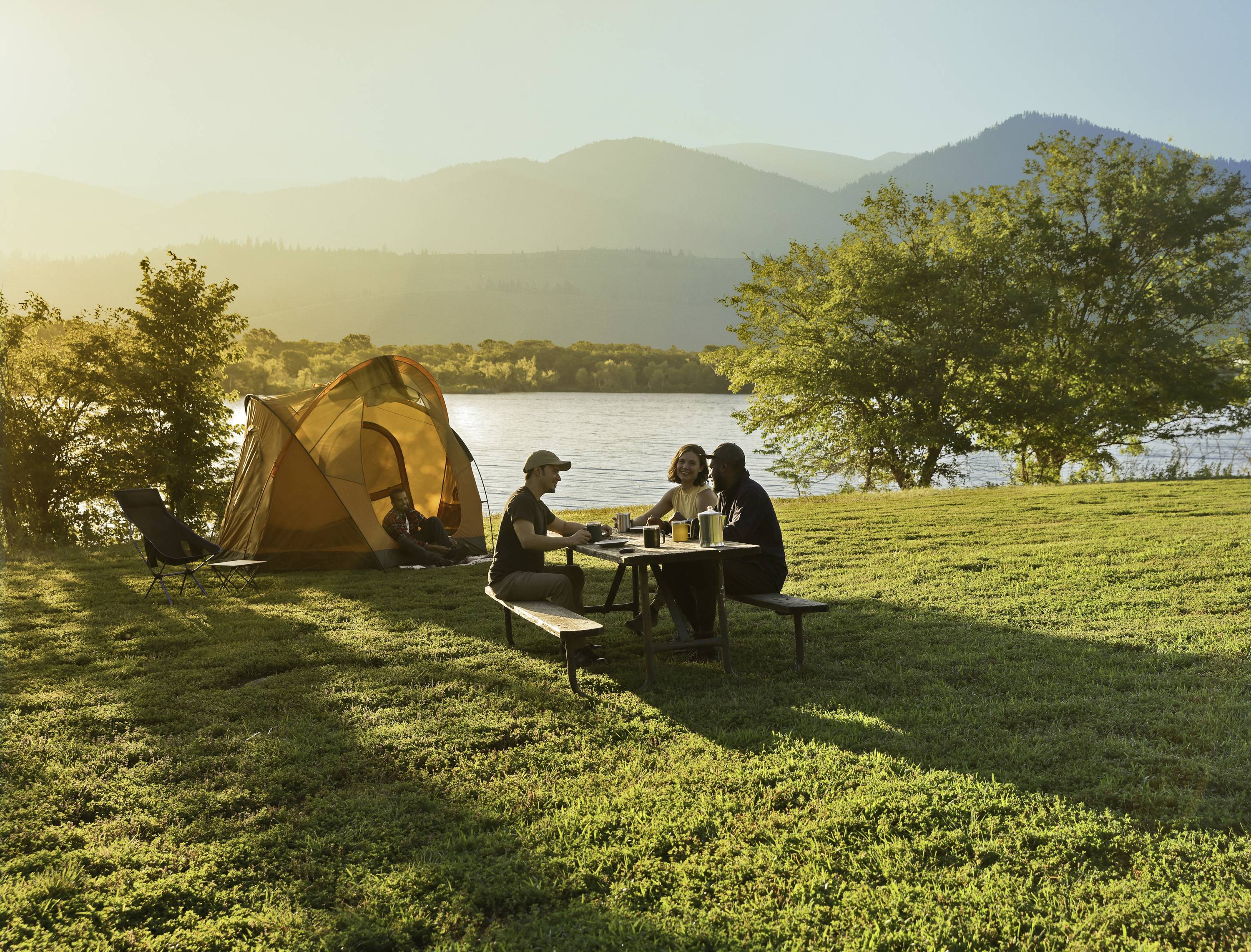 Drei Personen sitzen an einem Picknicktisch neben einem Zelt am See. Die Sonne geht unter, und im Hintergrund sind Berge zu sehen.
