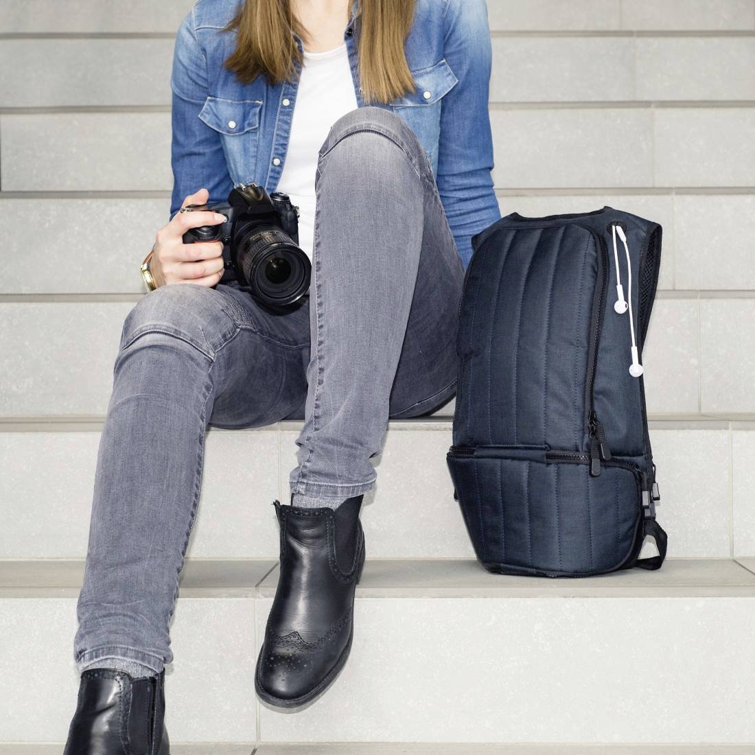 Eine Person sitzt auf einer Treppe, hält eine Kamera in der rechten Hand und hat einen Rucksack neben sich.