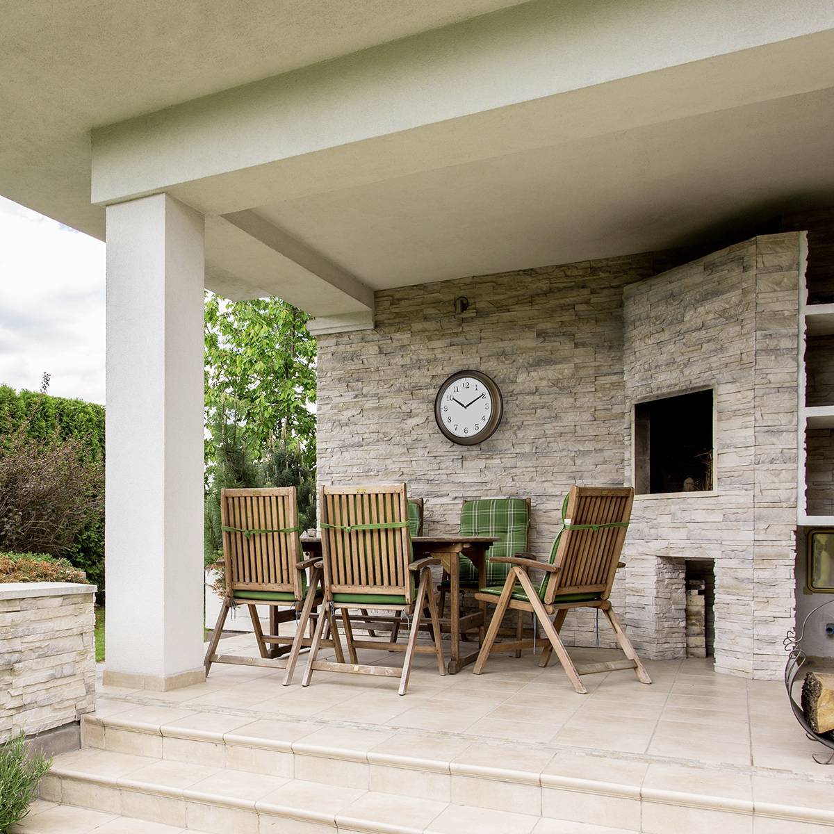 Eine Terrasse mit Tisch und Stühlen aus Holz. An der Wand hängt eine Uhr. Rechts ist ein eingebauter Grill zu sehen.