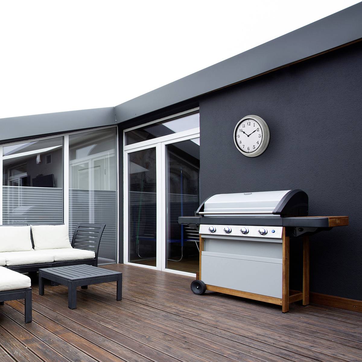 Terrasse mit Holzboden, weißem Sofa und modernem Grill vor einer grauen Hauswand mit runder Uhr.