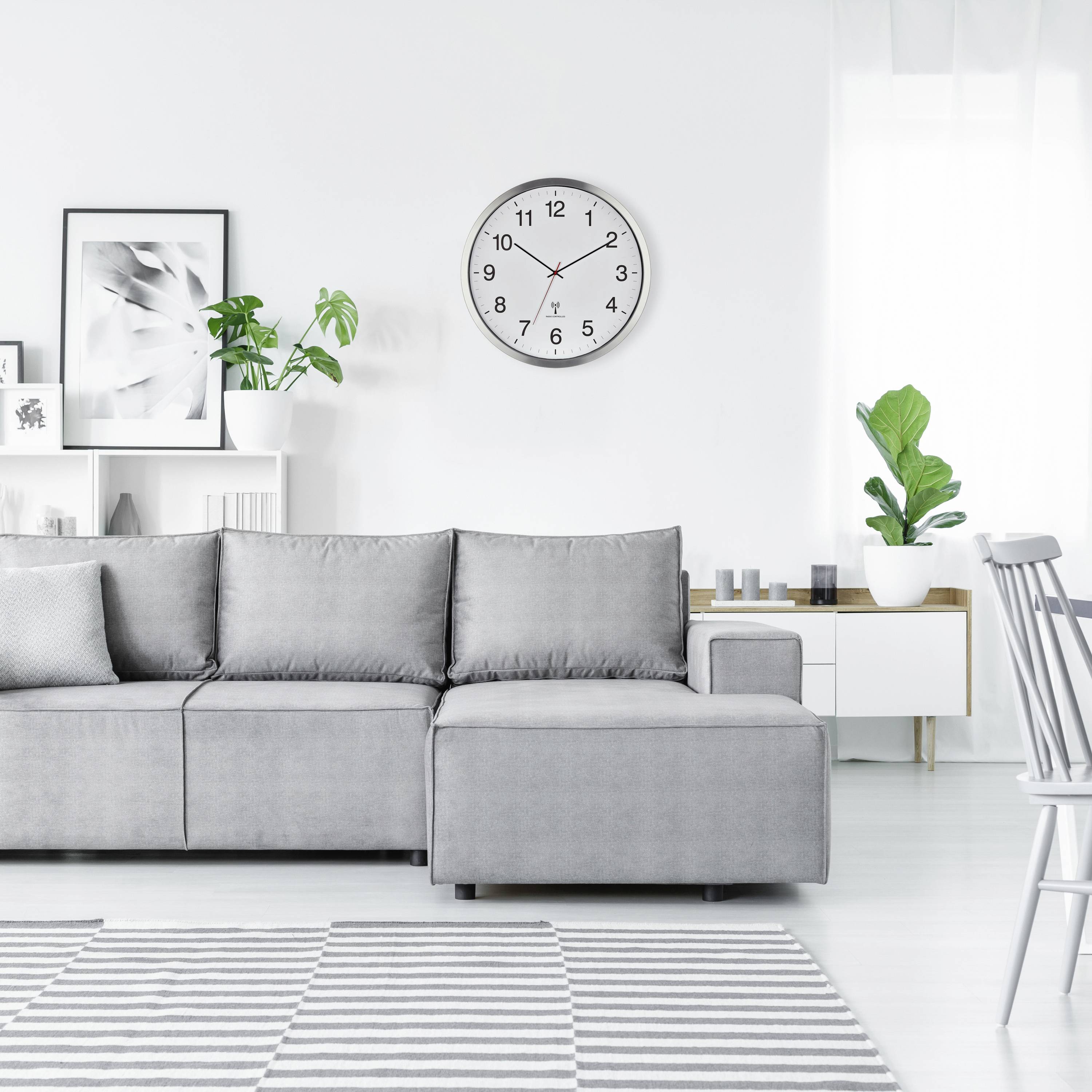 Graues Sofa in hellem Wohnzimmer mit Wandregalen, Pflanzen und einer runden Uhr. Moderner, minimalistischer Einrichtungsstil.