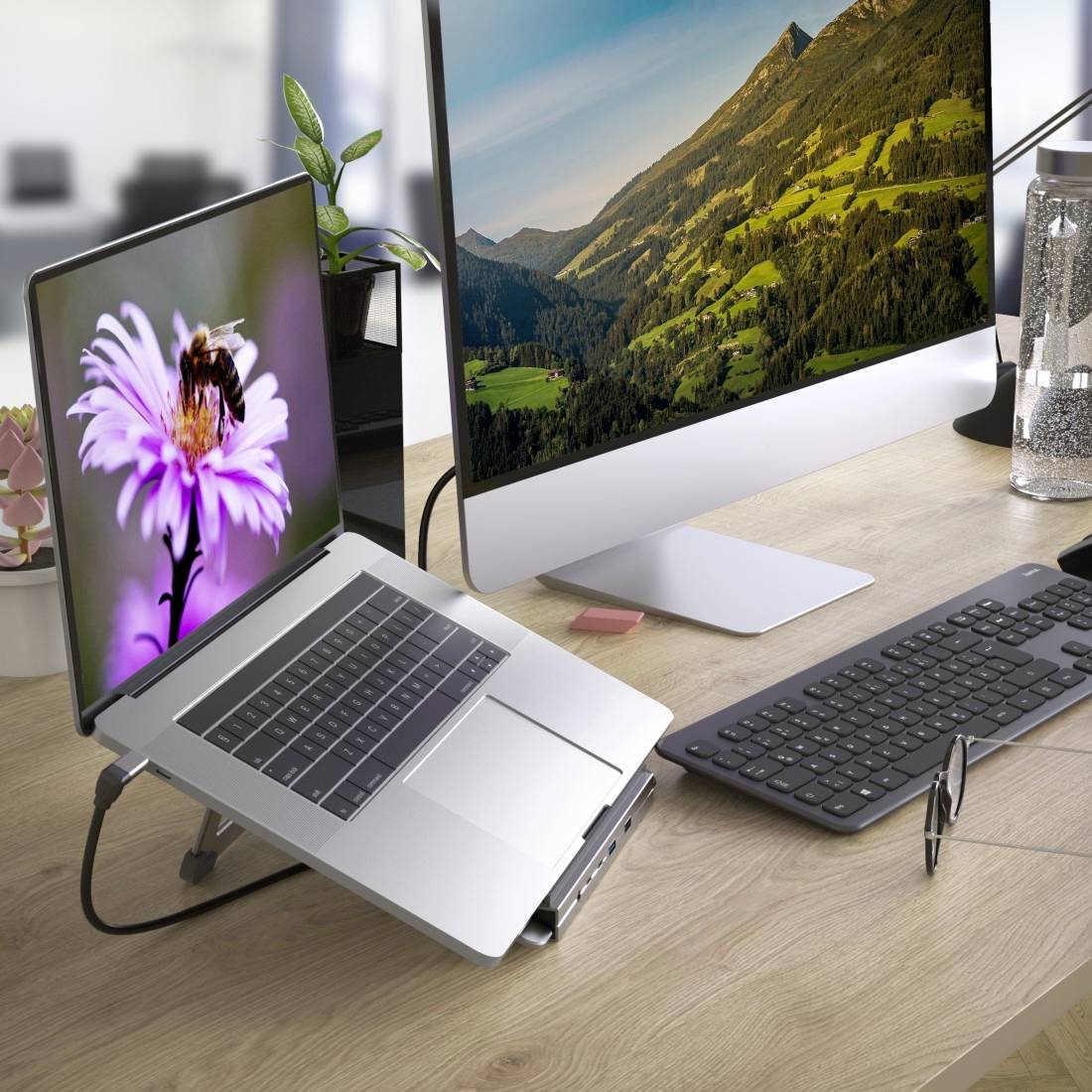 Ein Laptop mit Blumenbildschirm steht neben einem großen Desktop-Monitor mit Berglandschaft auf einem Schreibtisch mit Tastatur und Maus.