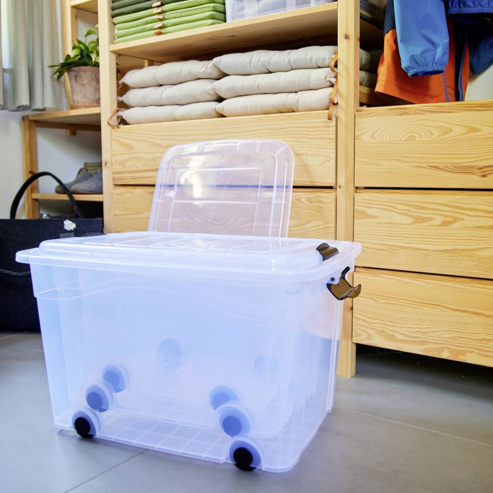 Eine transparente Plastikaufbewahrungsbox mit Rädern steht in einem Raum. Im Hintergrund sind Regale mit gefalteten Decken und Kleidung zu sehen.