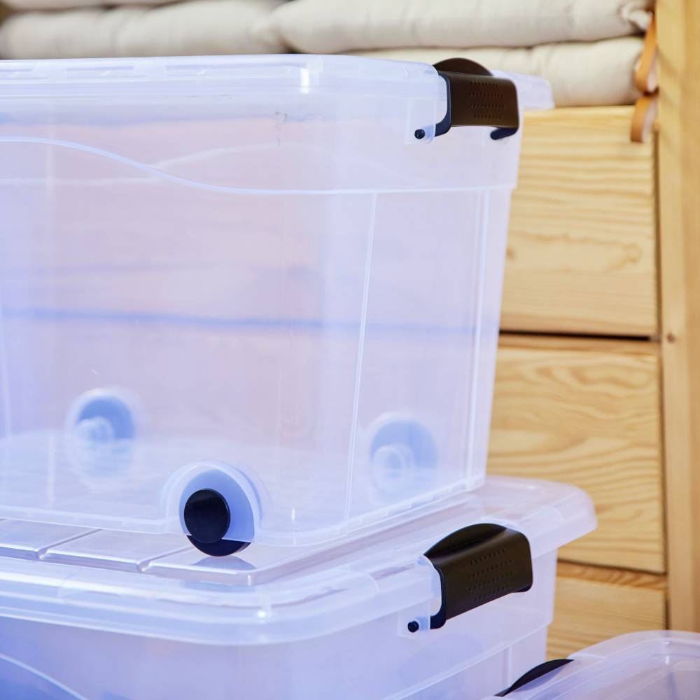 Durchsichtige Plastikboxen mit schwarzen Griffen stapeln sich. Im Hintergrund sind Holzmöbel sichtbar. Ideal zur Aufbewahrung von Gegenständen.