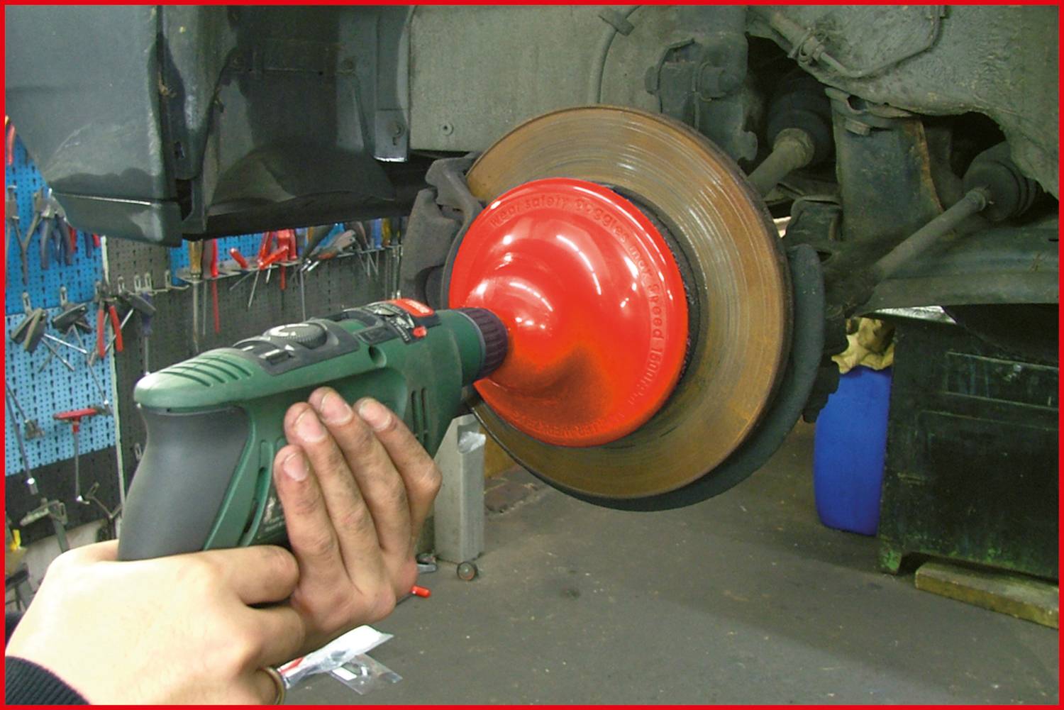 Eine Person reinigt mit einem Bohrmaschine-Adapter die Bremsscheibe eines Autos in einer Werkstatt. Im Hintergrund sind Werkzeuge sichtbar.