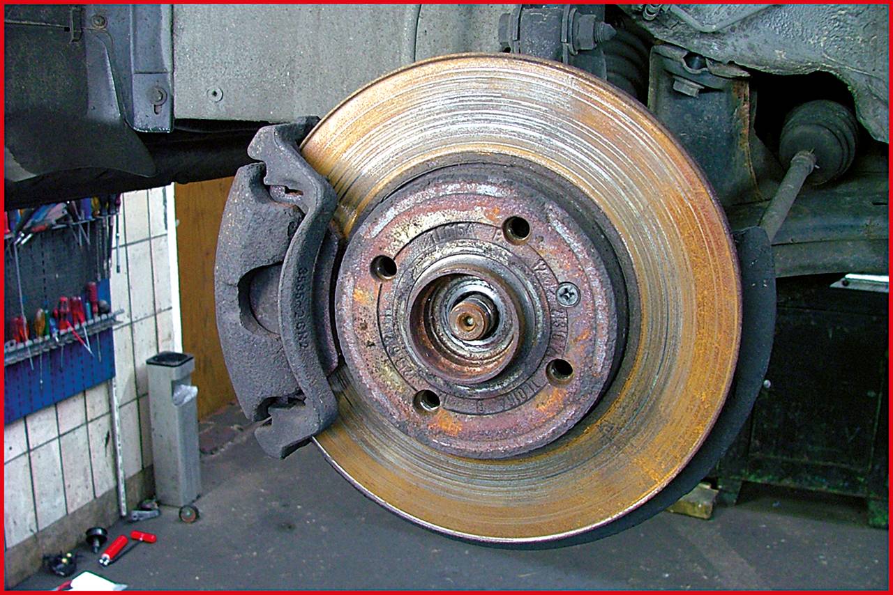 Nahaufnahme einer Autobremsanlage mit abgenutzter Bremsscheibe in einer Werkstatt. Werkzeuge und Teile sind im Hintergrund sichtbar.