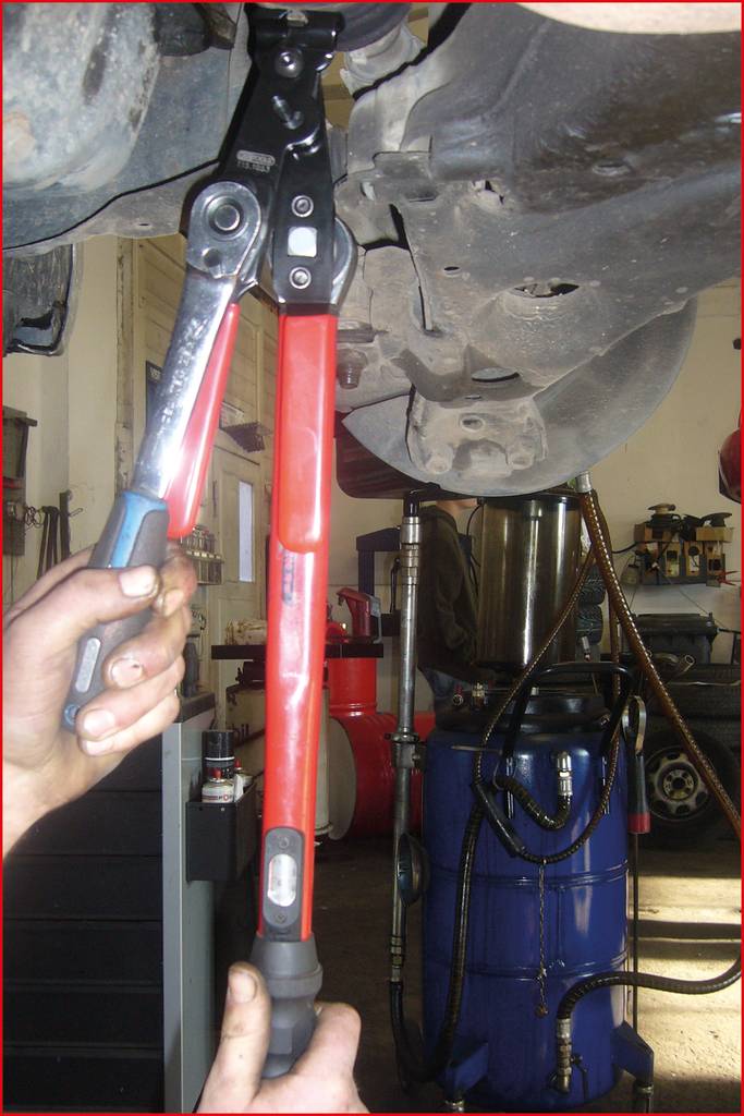 Eine Person benutzt einen langen, roten Hebel und einen Schraubenschlüssel, um an einem Fahrzeug zu arbeiten, das aufgebockt ist.