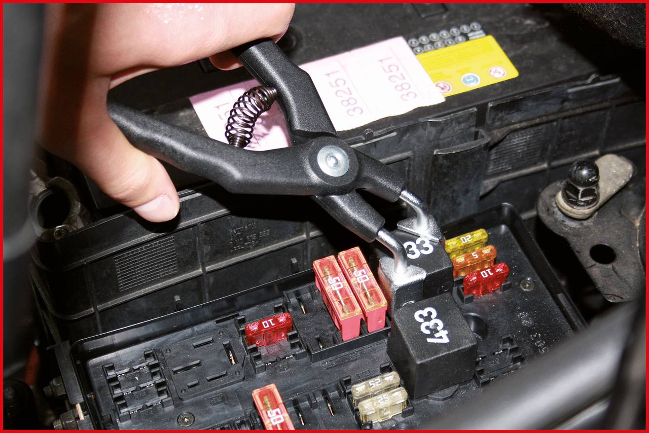 Eine Hand hält eine Klemme an einer Auto-Sicherung. Verschiedene farbige Sicherungen und Relais sind im Sicherungskasten zu sehen.