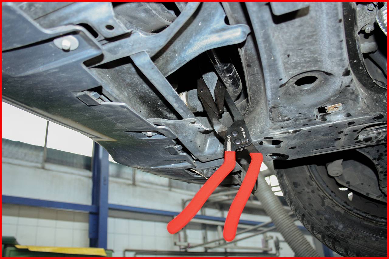 Zange klemmt unter einem Auto an einem Metallteil, in einer Werkstatt. Context des Werkzeugs bleibt unklar.
