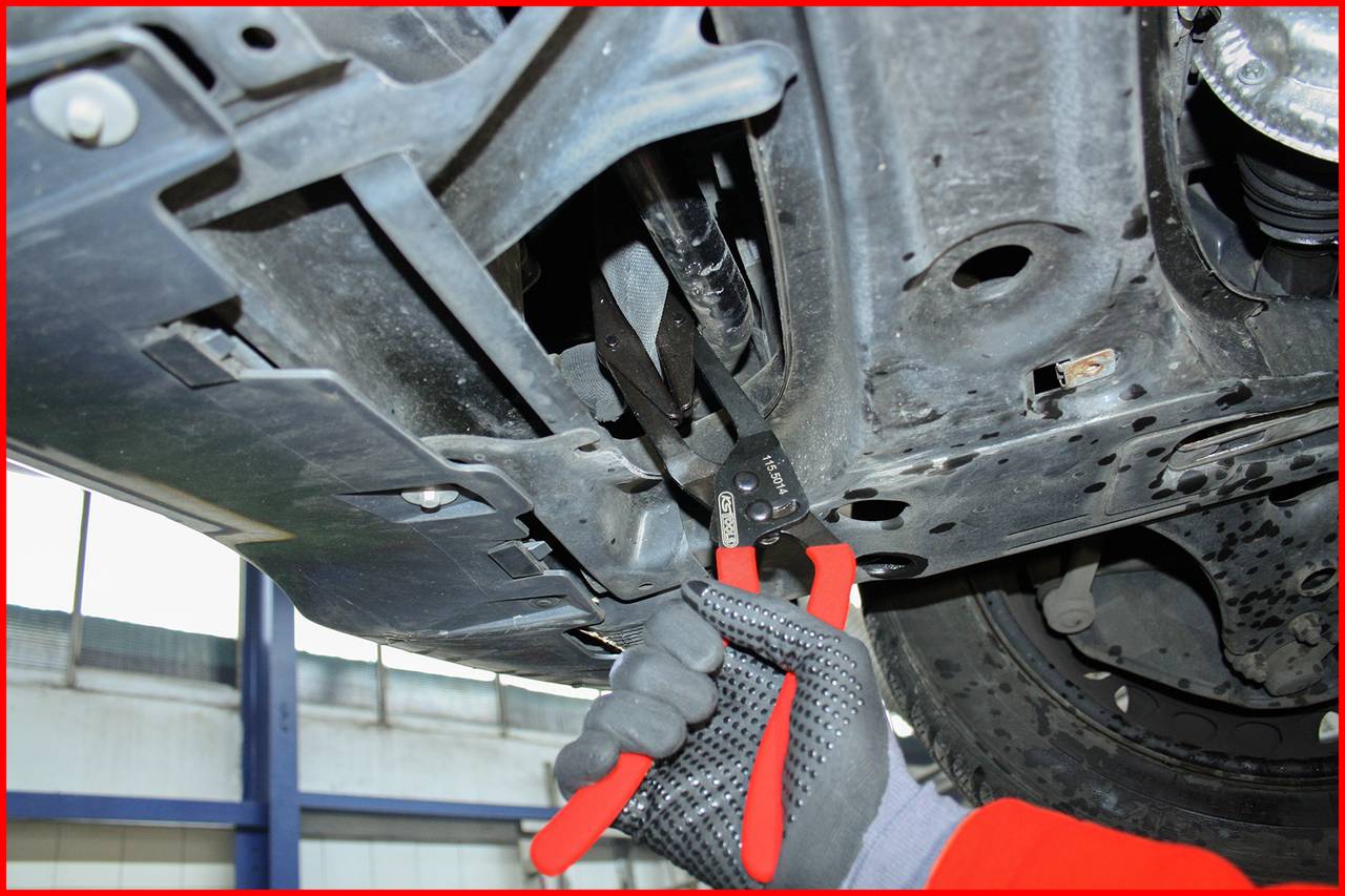 Mechanikerhand mit Zange zieht an einem Kabel unter einem Auto. Der Unterboden des Autos zeigt rostige Teile und Schrauben.