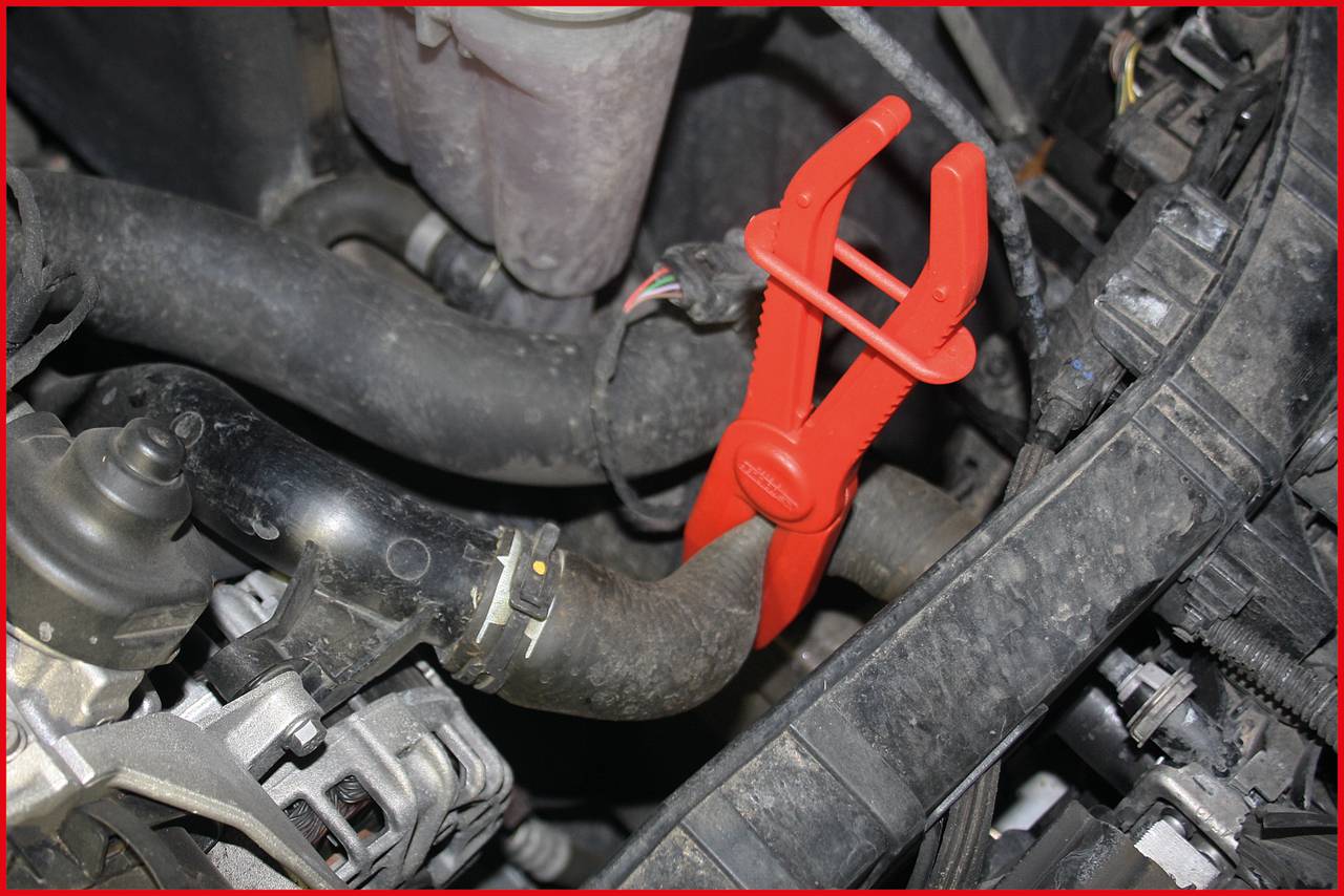 Ein roter Klemmknebel fixiert einen Gummischlauch im Motorraum eines Autos, um Wartungsarbeiten zu erleichtern.