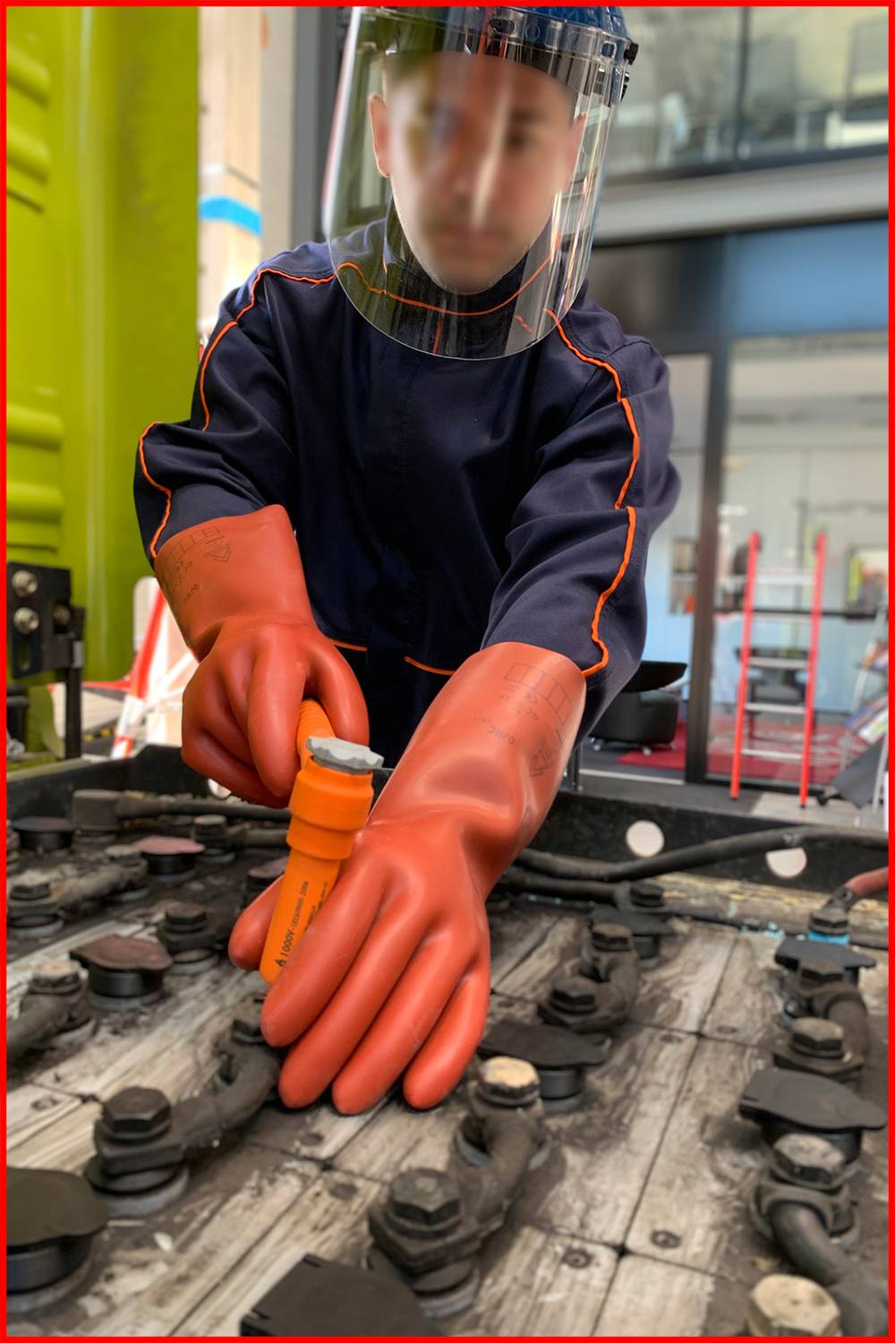 Eine Person in Schutzkleidung und Handschuhen arbeitet mit Werkzeugen an großen Batterien in einem industriellen Umfeld.