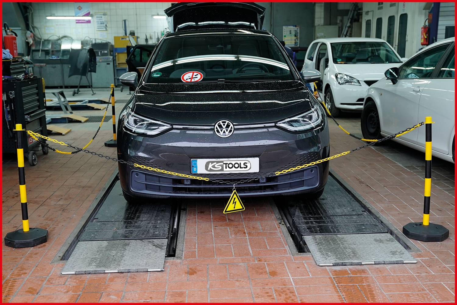 Ein graues Auto steht in einer Werkstatt auf einer Hebebühne. Es ist umgeben von Sicherheitsketten und anderen Fahrzeugen im Hintergrund.