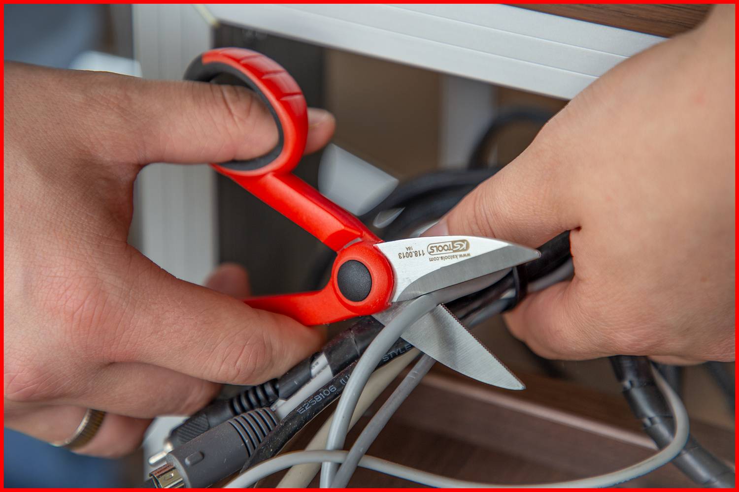 Zwei Hände halten eine rote Schere und schneiden mehrere schwarze und graue Kabel. Links ist ein Teil eines Möbelstücks zu sehen.