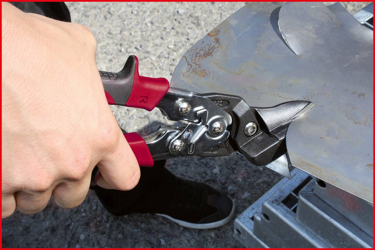 Hand hält rote Metallschere, die ein Stück Blech schneidet. Die Schere zeigt auf eine Kante des Metalls, Hintergrund ist unscharf.