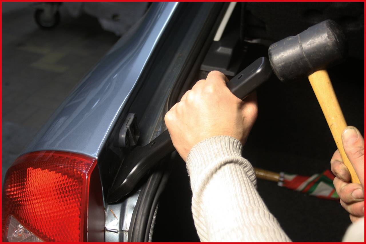 Eine Person benutzt einen Gummihammer und ein Werkzeug, um etwas an einem Autokofferraum zu reparieren oder einzupassen.