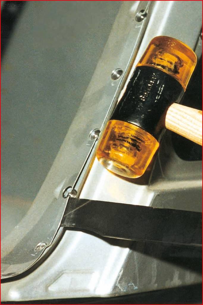 Ein Gummihammer drückt ein Metallstück an die Kante eines Autoteils und formt es. Die Szene zeigt einen Karosseriereparaturprozess.