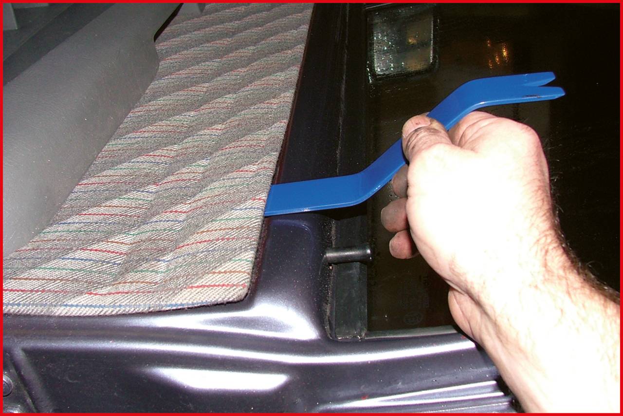 Hand verwendet ein blaues Hebelwerkzeug, um ein Autoteil zu entfernen. Teppichartige Abdeckung auf dem Armaturenbrett ist sichtbar.