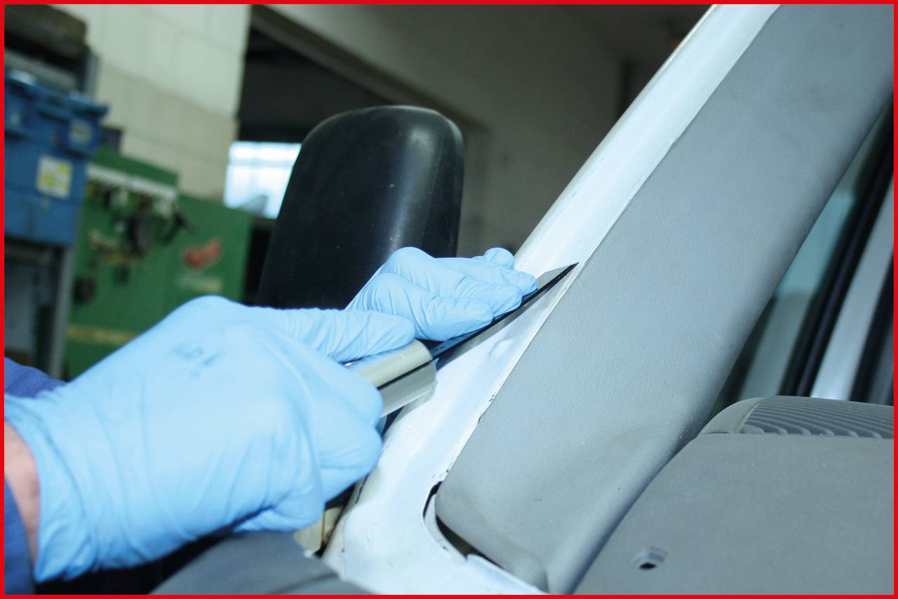 Zwei Hände mit blauen Handschuhen schneiden mit einem Messer Dichtmittel an einer Autoscheibe im Werkstattbereich ab.