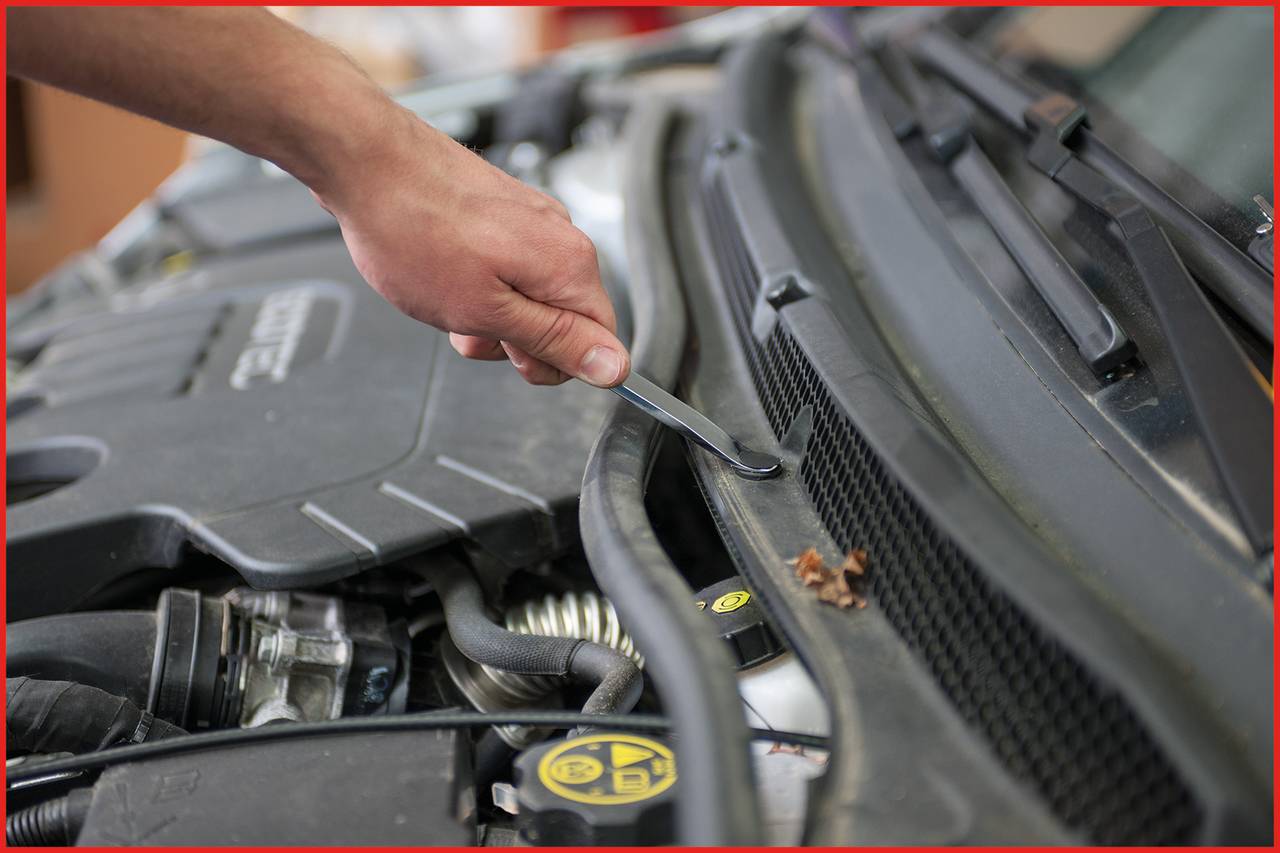 Eine Hand öffnet mit einem Schraubendreher die Motorhaube eines Autos. Mechanische Teile und der Markenname sind teilweise sichtbar.
