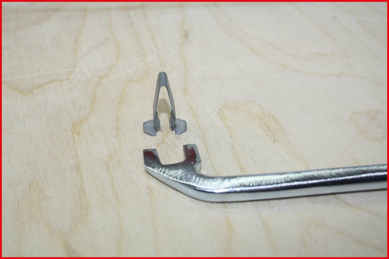 Ein Metallwerkzeug liegt auf einer Holzoberfläche, daneben ein kleines graues Bauteil. Das Werkzeug hat eine gebogene Form.