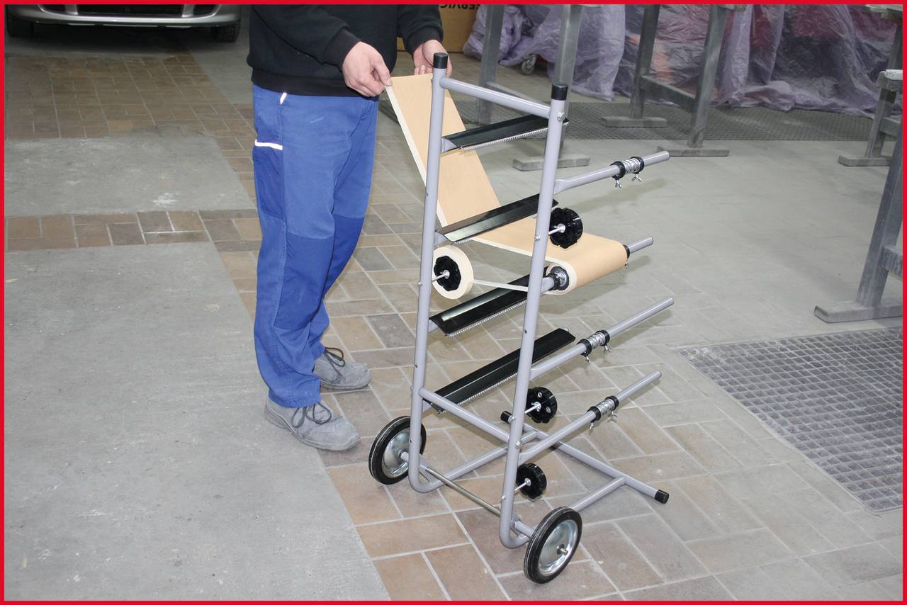 Eine Person steht neben einem mobilen Papierrollenhalter mit mehreren Rollen auf einem Industrie- oder Werkstattboden.