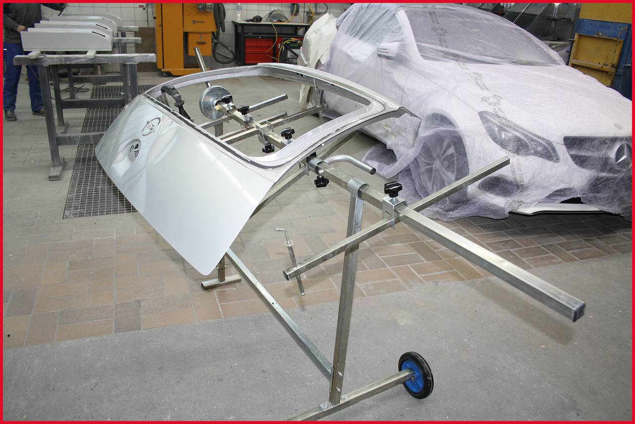Autogarage mit einer Autotür auf einem Gestell zur Reparatur. Im Hintergrund ein teilweise abgedecktes Auto. Werkzeuge und Geräte sind sichtbar.