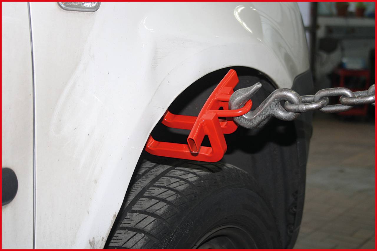 Ein weißes Auto wird mit einer roten Klammer an der vorderen Karosserie und einer Kette fixiert, möglicherweise in einer Werkstatt.