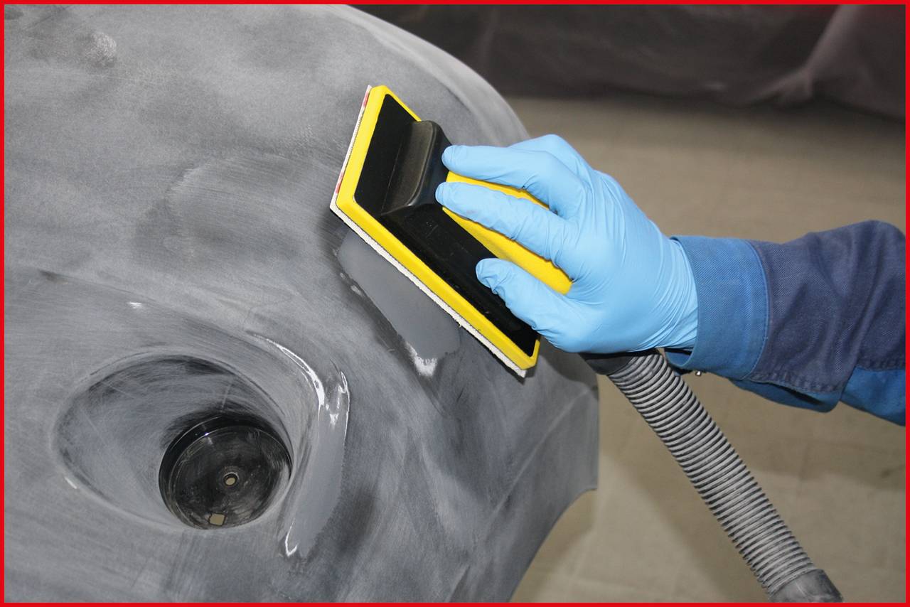 Person schleift ein Autoteil mit einem Handwerkzeug. Blaue Handschuhe und Staubabsaugschlauch sind sichtbar.