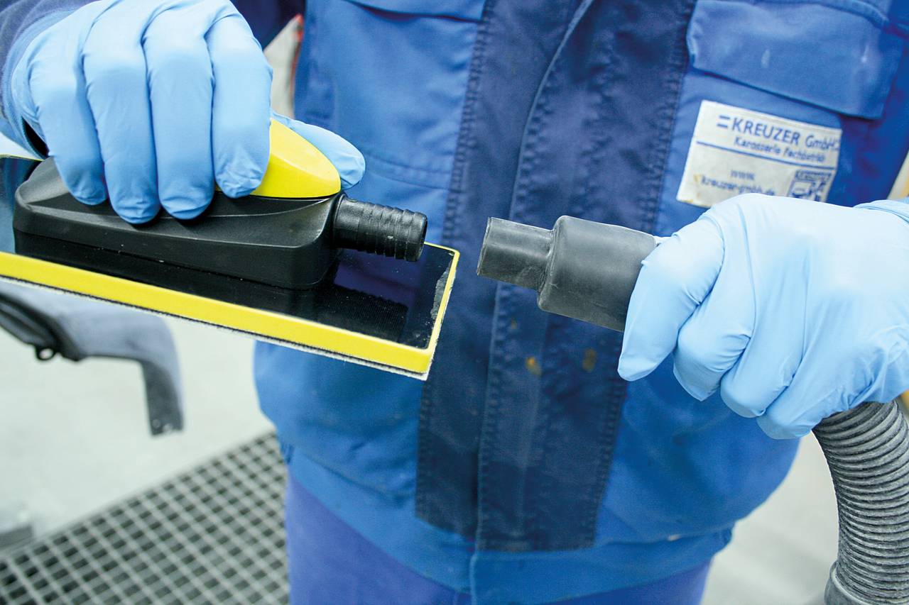 Person in blauer Arbeitskleidung hält ein Elektrowerkzeug und schließt es an einen Schlauch an. Arbeitsplatz in einer Werkstatt.