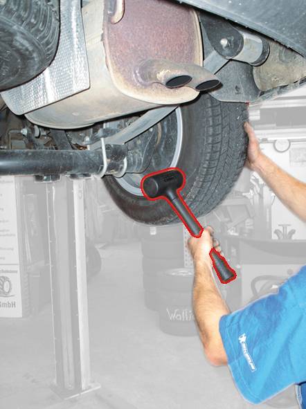 Person kontrolliert den Zustand eines Autoreifens, indem sie auf den Reifen klopft. Ein Hammer wird dabei verwendet.